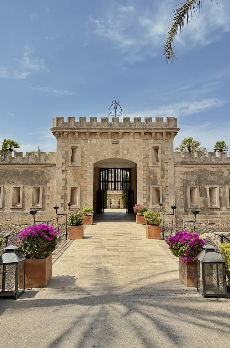 Historic stone fortress entrance at Cap Rocat, lined with magenta bougainvillea and palm trees under a bright Mallorca sky